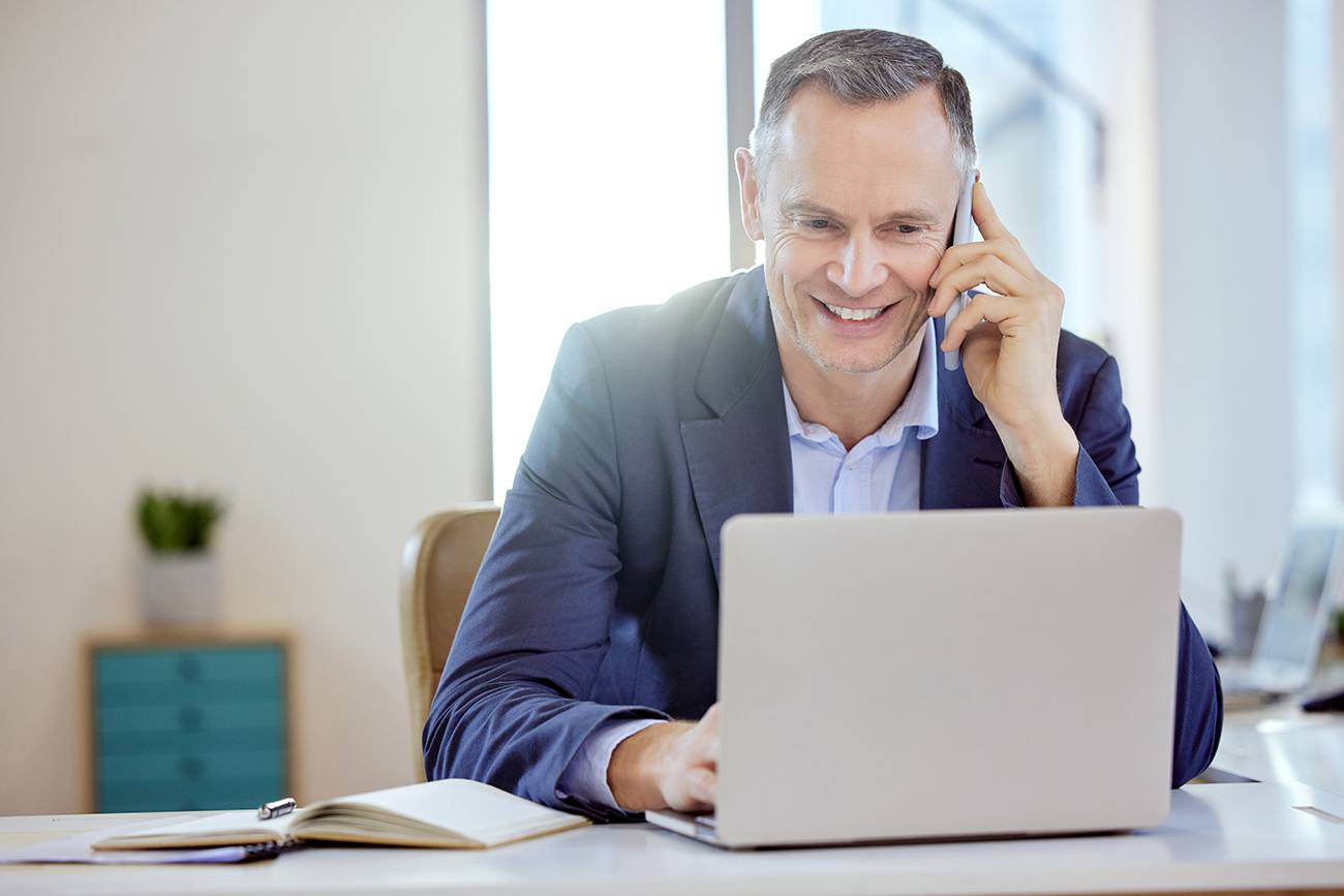 Muž v obleku sedí v kanceláři, telefonuje a pracuje na notebooku